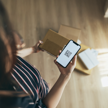 Eine Person hält ein Smartphone mit einem QR-Code in der Hand, während sie neben mehreren Kartons steht.