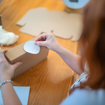 Eine Person hält ein braunes Paket mit einem Aufkleber in der Hand, während sie an einem Holztisch arbeitet.