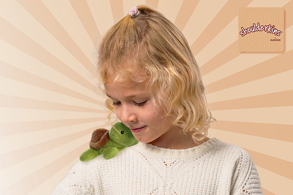 Ein Mädchen mit lockigem, blondem Haar trägt einen weißen Pullover und hat ein grünes Plüschschildkröten-Spielzeug auf der Schulter.
