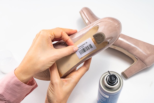 Eine Hand hält einen Schuh mit einem Preisschild, während eine Spraydose daneben steht. Der Schuh ist ein beiger Absatz.