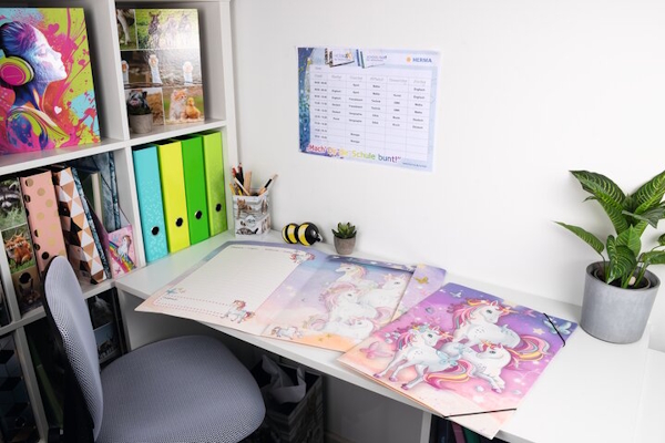 Ein Schreibtisch mit HERMA Sammelmappen aus der Einhorn Serie, einem Kalender an der Wand, bunten Ordnern und einer Pflanze in einem Topf.