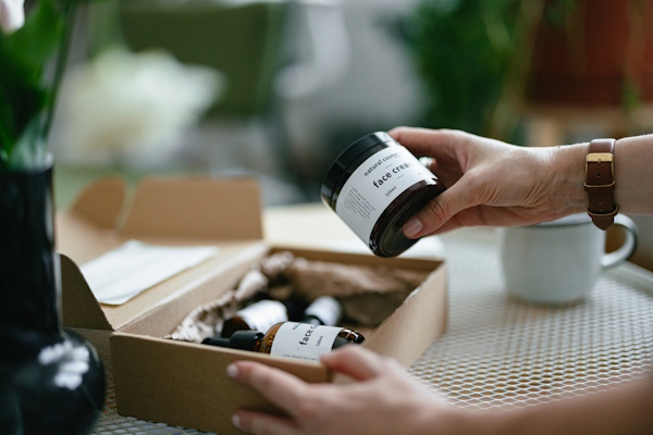 Eine Hand hält eine Kosmetikverpackung aus einem Karton heraus, die mit einem bedruckten Etikett versehen ist. In dem Karton befinden sich noch weitere Produkte, auf denen sich bedruckte Folienetiketten befinden.