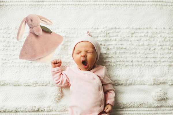 Ein schlafendes Baby in rosa Kleidung gähnt, während ein rosa Stoffhase daneben liegt. Das Baby trägt eine Mütze und liegt auf einer gestrickten Decke.