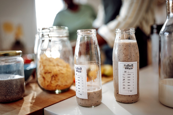 Zwei Flaschen mit Smoothies stehen auf einer Küchentheke, umgeben von verschiedenen Zutaten in Gläsern und Schalen.