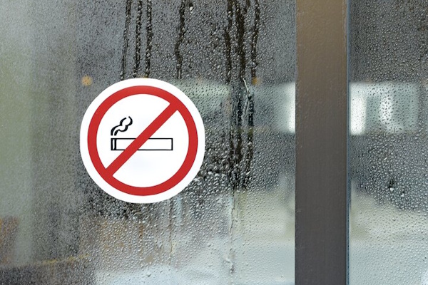 Ein rundes Schild mit einem durchgestrichenen Zigaretten-Symbol, das ein Rauchverbot anzeigt, ist an einer beschlagenen Glasfläche angebracht.