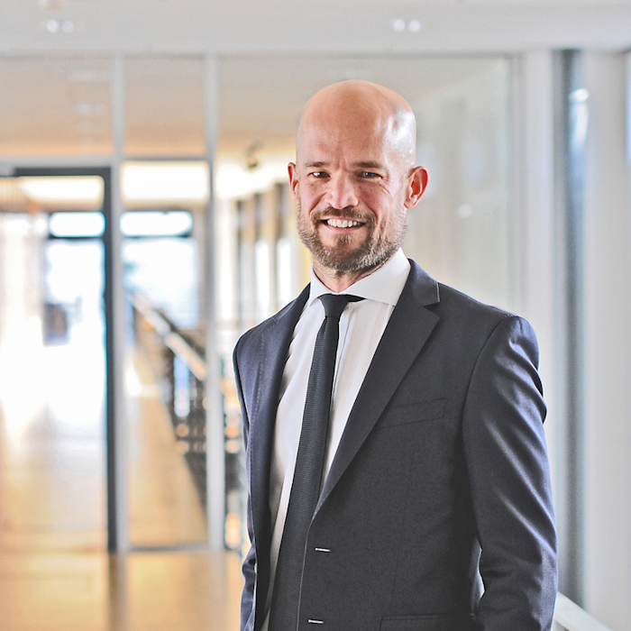 Ein Mann in einem Anzug steht in einem modernen Büroflur mit großen Fenstern. Er lächelt und trägt eine Krawatte.