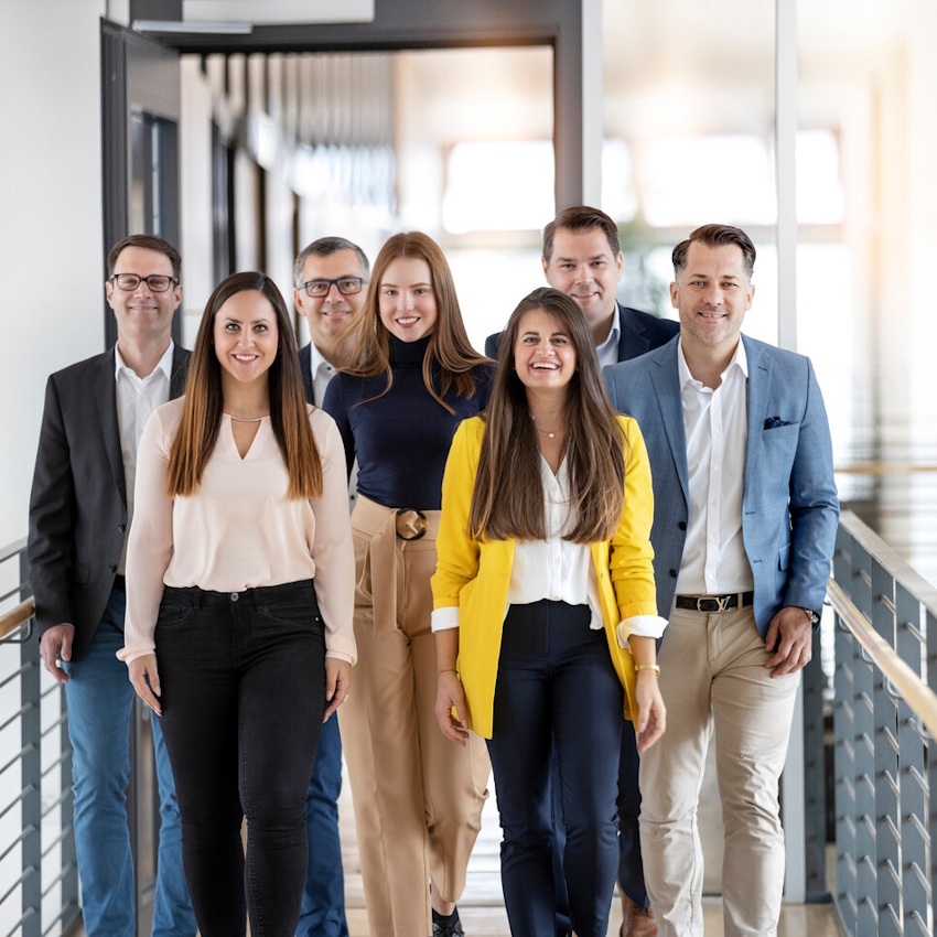 Eine Gruppe von sieben Personen steht in einem Flur, lächelnd und in formeller Kleidung, bereit für ein Treffen oder eine Veranstaltung.