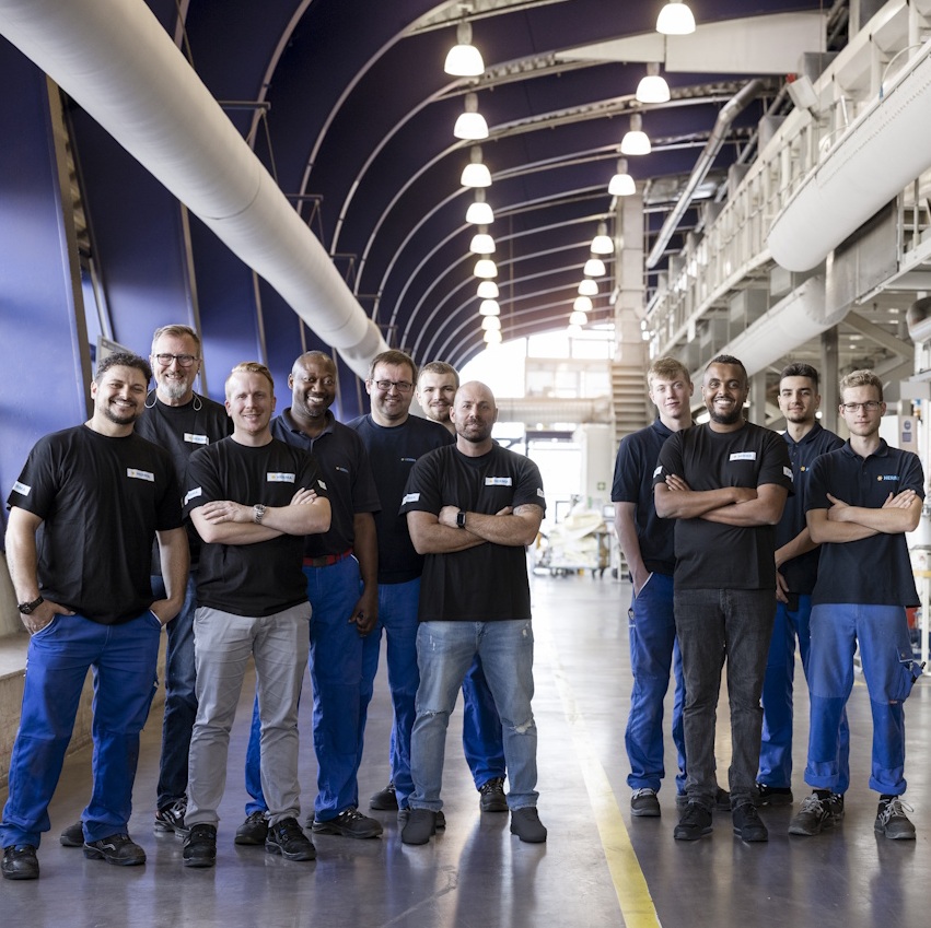 Eine Gruppe von zehn Personen in Arbeitskleidung steht in einer Fabrikhalle, lächelnd und mit verschränkten Armen, vor einer industriellen Kulisse.