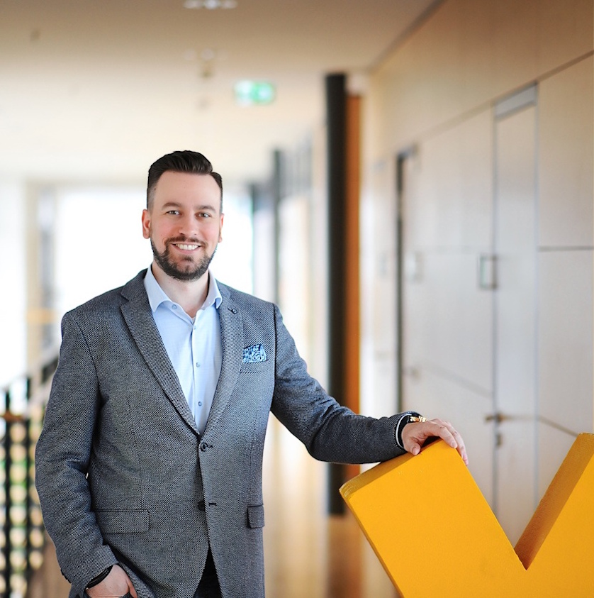 Mann in Anzug steht in einem modernen Flur neben einem großen gelben Buchstaben. Er lächelt und trägt eine blaue Hemd mit einer passenden Tasche.