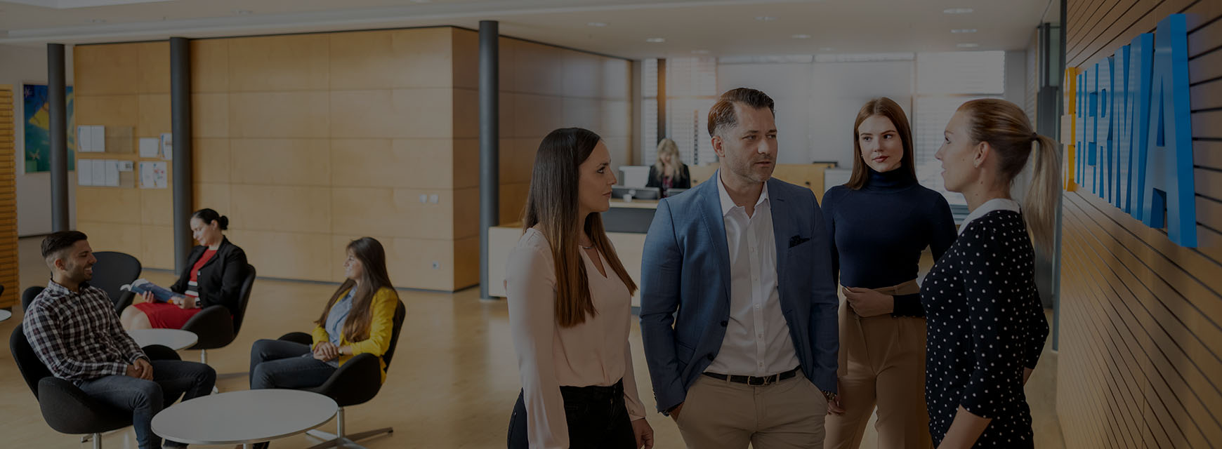 Fünf Personen in einem modernen Büro sprechen miteinander, während zwei weitere Personen in der Hintergrund entspannt sitzen.