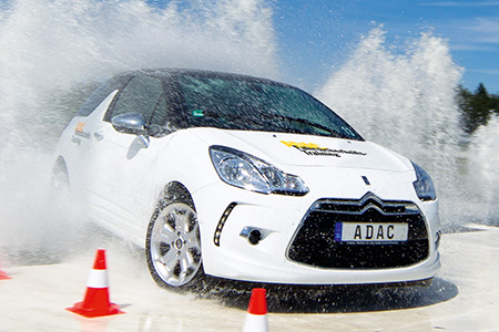 Ein weißer Citroën fährt durch Wasser, spritzt Wasser auf, umgeben von Pylonen. ADAC-Logo sichtbar.