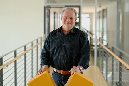 Ein Mann steht in einem Flur und hält zwei gelbe Objekte in den Händen. Er trägt ein schwarzes Hemd und lächelt leicht.