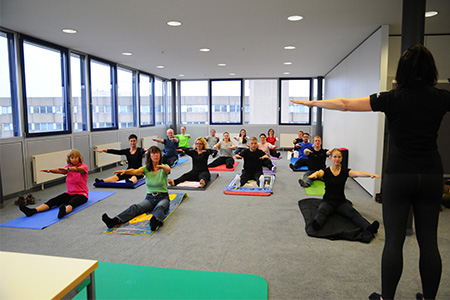 Eine Gruppe von Menschen führt Übungen in einem Raum durch, während eine Trainerin Anweisungen gibt. Matten und Fitnessgeräte sind vorhanden.