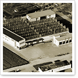 Luftaufnahme eines großen Gebäudes mit mehreren Dächern, umgeben von Straßen und Feldern. Das Gebäude scheint eine industrielle Nutzung zu haben.