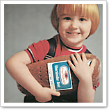 Ein lächelndes Kind mit blonden Haaren hält einen Football und trägt ein rotes T-Shirt mit einem schwarzen Rucksack.