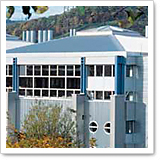 Moderne Gebäude mit großen Fenstern und blauen Akzenten, umgeben von Bäumen und Hügeln im Hintergrund.