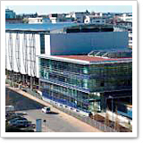 Moderne Gebäude mit Glasfassade und flachem Dach, umgeben von einer Straße und weiteren urbanen Strukturen.