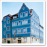 Blaues Gebäude mit mehreren Fenstern und einem Eingang, beschriftet mit "HITRIVA". Das Dach hat eine markante Form.