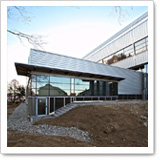 Moderne Gebäude mit Glasfassade und schrägem Dach, umgeben von einem unbefestigten Bereich und Bäumen im Hintergrund.
