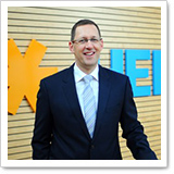 Ein Mann in einem Anzug steht vor einer Holzwand mit einem orangefarbenen und blauen Logo. Er lächelt und wirkt professionell.