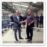 Zwei Männer schütteln sich die Hände in einer Fabrikhalle, während mehrere Personen im Hintergrund stehen.
