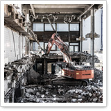 Bagger arbeitet in einem abgerissenen Gebäude, umgeben von Trümmern und Baustellenmaterial. Fenster und Wände sind teilweise entfernt.