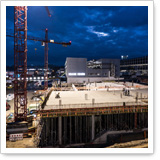 Bauplatz mit Betonfundament und Kränen unter bewölktem Himmel. Baustellenaktivitäten sind sichtbar, mit Gebäudestrukturen im Hintergrund.