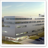 Moderne, mehrstöckige Gebäude mit großen Fenstern und einer hellen Fassade, umgeben von einer grünen Landschaft und parkenden Autos.