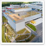 Luftaufnahme eines modernen Gebäudes mit begrüntem Dach, umgeben von Wegen und Grünflächen, in einem industriellen Umfeld.