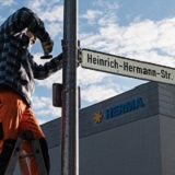 Ein Arbeiter steht auf einer Leiter und montiert ein Straßenschild mit der Aufschrift "Heinrich-Hermann-Str." vor einem Firmengebäude.