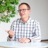 Ein Mann mit Brille und kariertem Hemd sitzt an einem Tisch und gestikuliert, während er spricht. Im Hintergrund ist eine Pflanze sichtbar.
