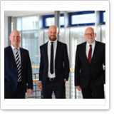 Drei Männer in Anzügen stehen in einem modernen Bürogebäude. Sie lächeln und posieren freundlich vor großen Fenstern.