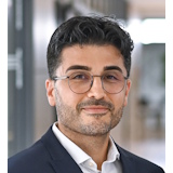 Ein Mann mit dunklen, lockigen Haaren und Brille, trägt einen Anzug und steht in einem modernen Innenraum.