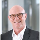 Ein Mann mit Brille und Glatze trägt einen Anzug und lächelt freundlich. Der Hintergrund ist unscharf und hell.