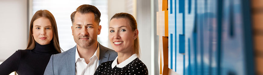 Drei Personen stehen in einem modernen Büro. Der Mann in einem Anzug und zwei Frauen, eine in einem schwarzen Rollkragenpullover und die andere in einem gepunkteten Oberteil.