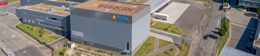 Moderne Lagerhalle mit begrüntem Dach und dem Logo von "Hertila" an der Fassade, umgeben von Parkplätzen und Straßen.