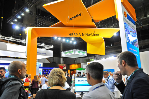 Menschen betrachten den HERMA InNo-Liner Stand auf einer Messe, umgeben von verschiedenen Ausstellungsständen und Beleuchtung.