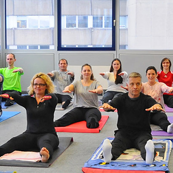 Eine Gruppe von Menschen praktiziert Yoga in einem Raum, einige halten ihre Arme seitlich ausgestreckt, während andere auf Matten sitzen.