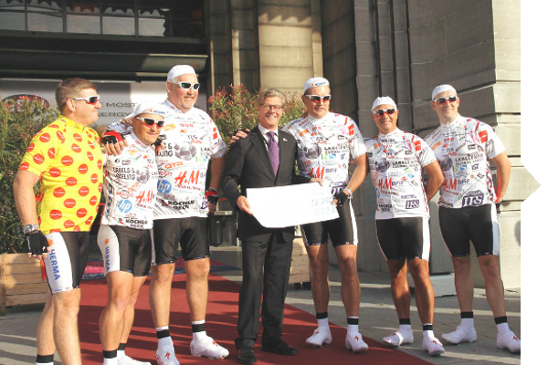 Sechs Radfahrer in Trikots stehen mit einem Mann in Anzug und Krawatte, der ein großes Schild hält, vor einem Gebäude.