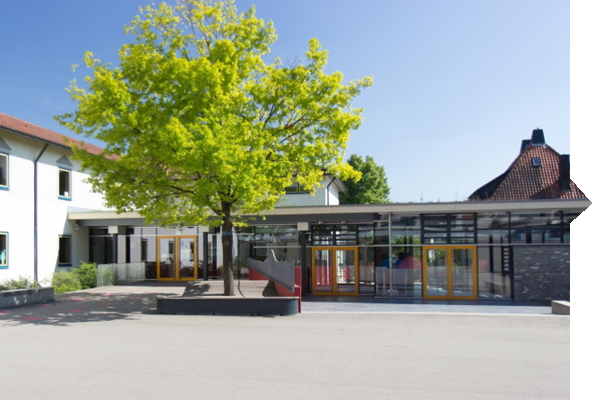 Ein Schulgebäude mit modernen Fenstern, einem großen Baum im Vordergrund und einem Spielbereich, der durch farbige Türen zugänglich ist.