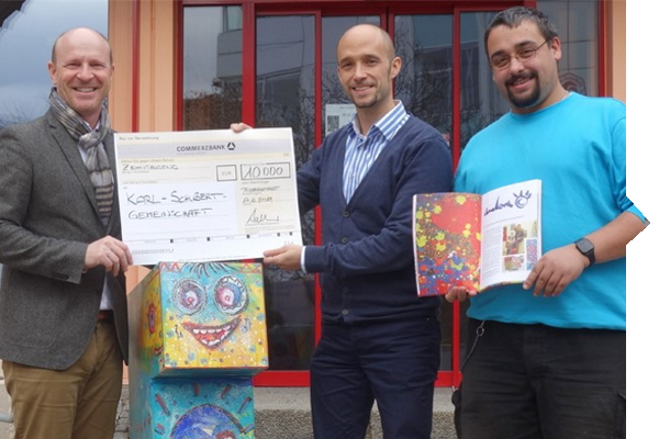Drei Männer halten einen großen Scheck über 10.000 Euro und ein buntes Kunstbuch. Im Hintergrund ist ein farbenfrohes Kunstwerk zu sehen.