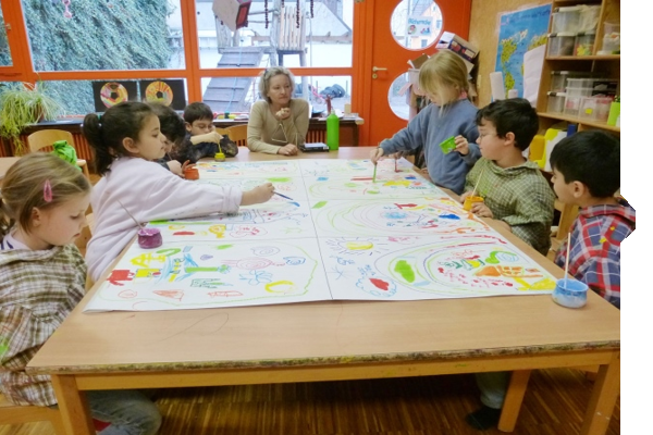 Kinder malen gemeinsam an einem großen Tisch, während eine Erwachsene sie anleitet. Bunte Zeichnungen und Malutensilien sind sichtbar.