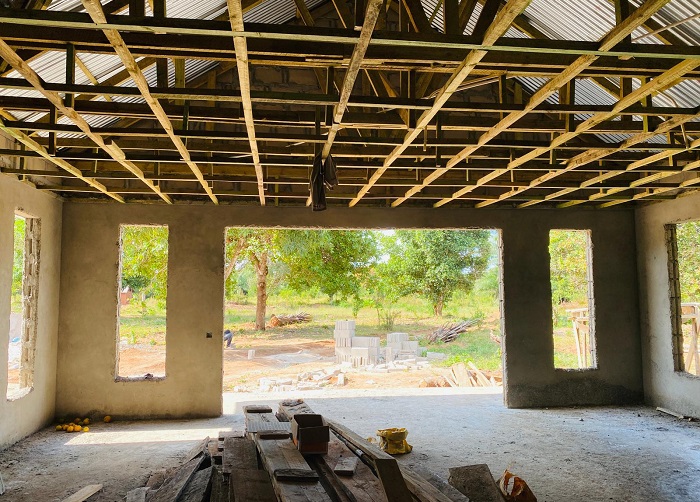 Unvollendeter Raum mit freiliegendem Dachstuhl und großen Fenstern, die auf einen grünen Außenbereich blicken. Holzmaterialien liegen auf dem Boden.