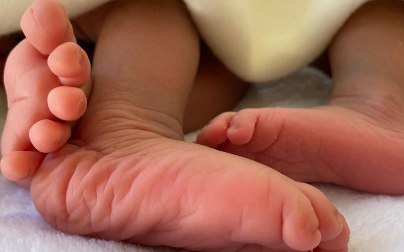 Zwei kleine Babyfüße liegen auf einer weichen Unterlage, teilweise von einer Decke bedeckt. Die Haut ist zart und rosig.