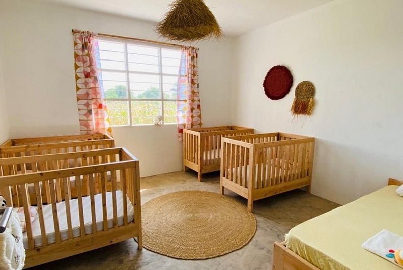 Ein heller Raum mit vier Holz-Kinderbetten, einem runden Teppich und bunten Vorhängen. Ein Fenster bietet Blick nach draußen.