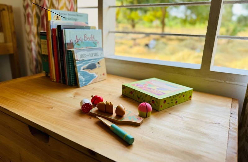 Bunte Holzspielzeuge und ein Spielzeugmesser liegen auf einem Holztisch, während Bücher im Hintergrund stehen.