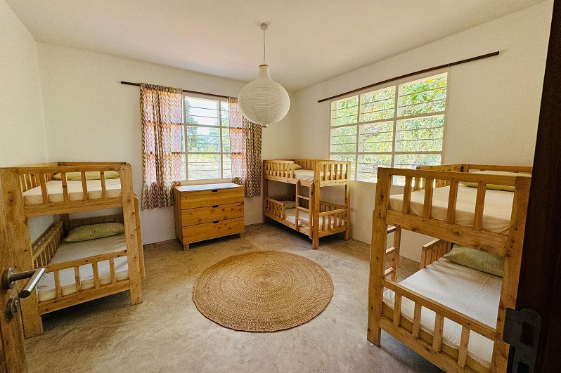 Ein helles Kinderzimmer mit zwei Etagenbetten aus Holz, einem runden Teppich und einem Kommode. Fenster mit Vorhängen sorgen für Tageslicht.