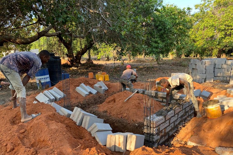 Arbeiter bauen mit Ziegeln und Beton in einem Baustellenbereich, umgeben von Erde und Baumvegetation.