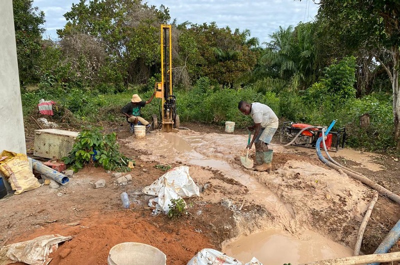 Zwei Arbeiter bohren ein Loch in den Boden, während eine Bohrmaschine im Hintergrund steht. Umgeben von Erde und Wasser.