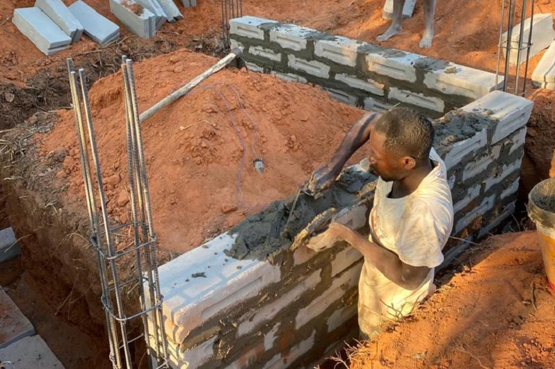 Ein Arbeiter mauert mit Zementsteinen an einer Baustelle, um eine Wand zu errichten. Der Boden ist mit rotem Sand bedeckt.